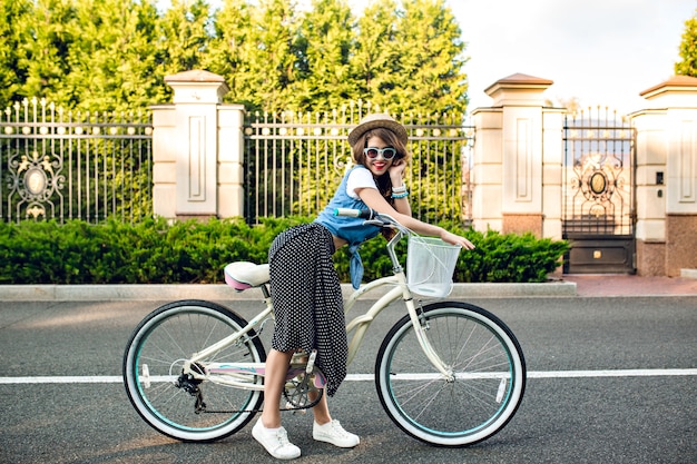 Attraktives Mädchen mit dem langen lockigen Haar im Hut, das mit Fahrrad auf Straße aufwirft. Sie trägt einen langen Rock, ein Wams und eine blaue Sonnenbrille. Sie lächelt in die Kamera.