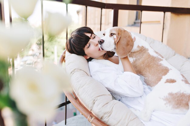 Attraktives Mädchen mit dunklen kurzen Haaren, die mit Liebe Beagle Hund küssen, der wegschaut