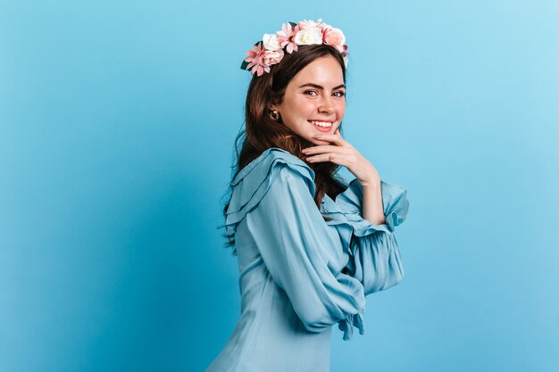 Attraktives Mädchen mit schelmischem Lächeln schaut in die Kamera. Foto der Dame im blauen Kleid mit Blumenkrone.