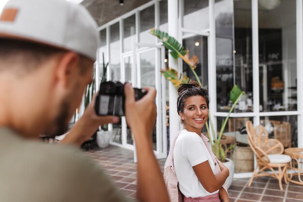 Attraktives Mädchen mit stilvollem Brötchen lächelt, während ihre Freundin Fotos macht Porträt einer Frau in weißem T-Shirt und rosa Jacke