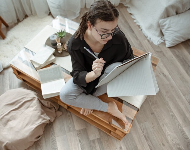 Attraktives Mädchen zu Hause, das mit einem Notizbuch und Stift unter den Büchern Draufsicht sitzt.