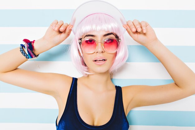 Attraktives Modell des attraktiven Sommerporträts der Nahaufnahme im blauen Badeanzug mit geschnittener rosa Frisur, rosa Sonnenbrille Lookong auf gestreifter blau-weißer Wand. Junge sexy Frau, erstaunliche, fröhliche Stimmung.