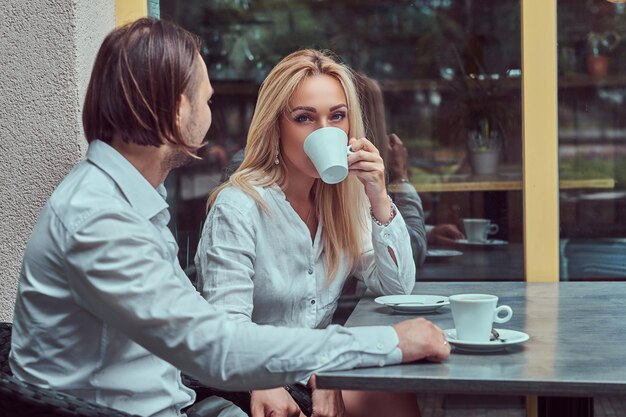 Attraktives Paar - charmante blonde Frau in weißer Bluse und bärtiger Mann mit stylischem Haarschnitt in weißem Hemd bei einem Date in einem Café im Freien.