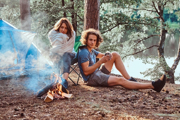 Kostenloses Foto attraktives paar junger leute, die am lagerfeuer im grünen, sonnigen wald chillen. sie haben ein blaues zelt.