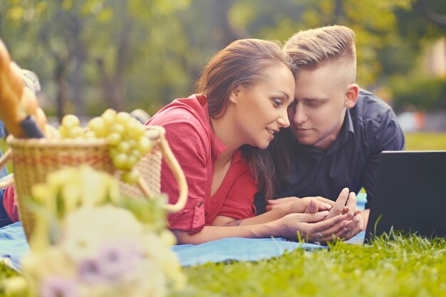Attraktives Paar liegt auf einer Decke auf einem Rasen und benutzt einen Laptop für ein Picknick.
