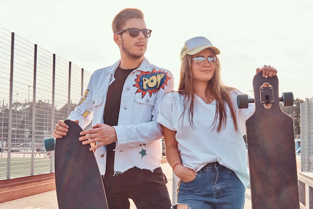 Attraktives Paar trendig gekleideter junger Hipster, die an sonnigen Tagen mit Skateboards im städtischen Sportkomplex posieren, mit warmen Tönen.