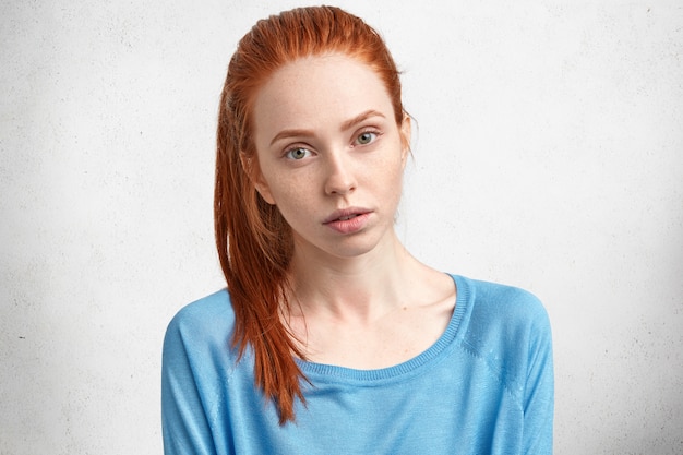 Kostenloses Foto attraktives rothaariges weibliches model mit ernstem, selbstbewusstem ausdruck, gesunder sommersprossiger haut, hellblauem pullover und posen an der weißen betonwand.