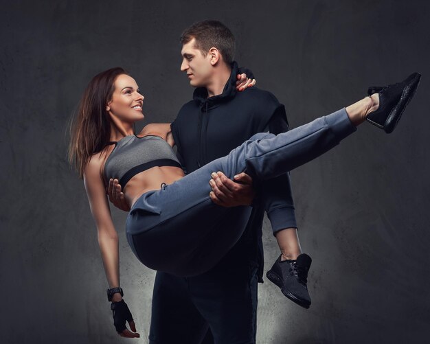 Attraktives Sportpaar. Schöner Kerl, der seine Freundin auf seinen Händen hält und Sportkleidung auf einem dunklen, strukturierten Hintergrund trägt.