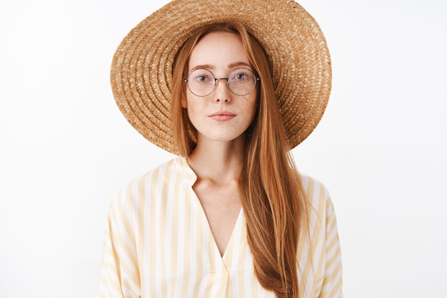 Attraktives stilvolles Hipster-Mädchen mit Ingwerhaar und Sommersprossen im trendigen Strohhut der Brille und gelber niedlicher Bluse, die mit erfreutem sorglosem Ausdruck lächeln, der interessanten Vortrag im Café besucht