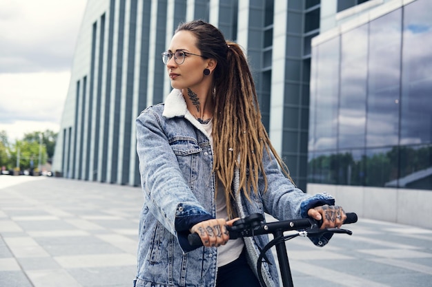 Attraktives stylisches Mädchen mit Dreadlocks fährt Elektroroller auf der Straße in der Nähe eines interessanten Gebäudes.