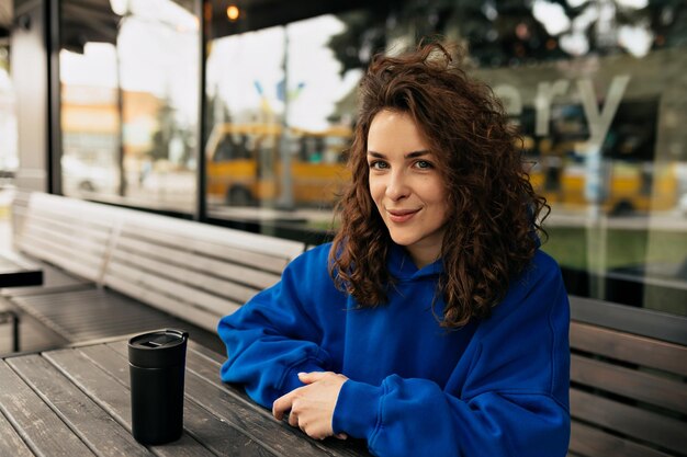 Attraktives süßes Mädchen mit üppiger Frisur in blauem, stilvollem Pullover posiert vor der Kamera im Freien, während es sich auf der Sommerterrasse mit Kaffee ausruht