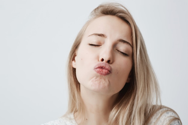 Attraktives verträumtes weibliches Model mit blonden Haaren schließt Augen, schmollt Lippen, schickt Küsse