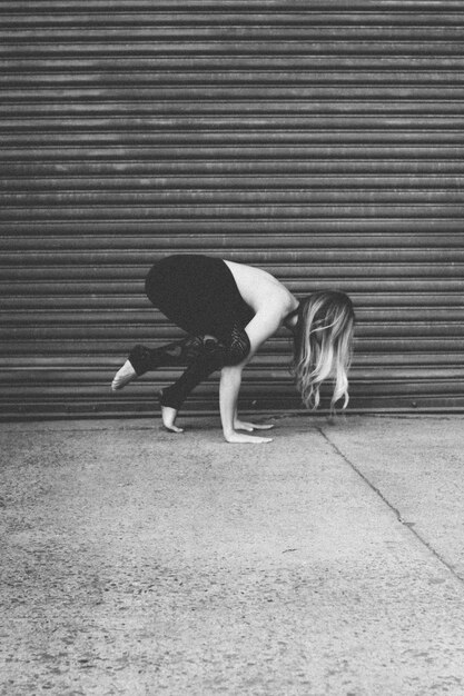 Attraktives weibliches Model, das Yoga in der Nähe einer Garage auf dem Bürgersteig praktiziert, in Graustufen aufgenommen