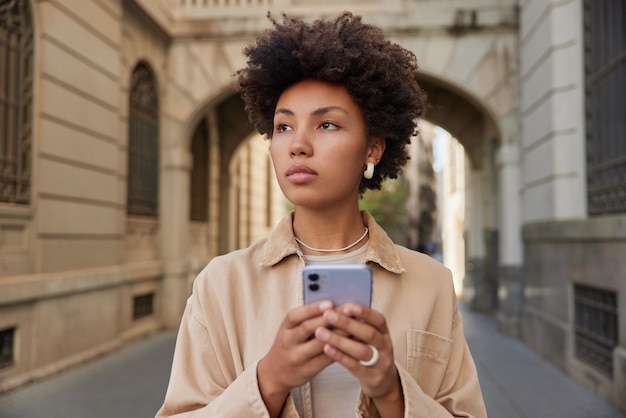 Attraktives weibliches Modell liest Textnachricht auf Smartphone-Spaziergängen während des Rundgangs im Freien hat nachdenklichen Ausdruck genießt Erholungszeit Posen in der Nähe des alten Gebäudes Junger Tourist mit Telefon