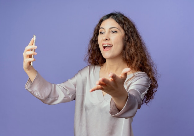 Auf der Seite freudiges junges hübsches Mädchen, das Telefon hält und Hand lokalisiert auf blauer Wand hält