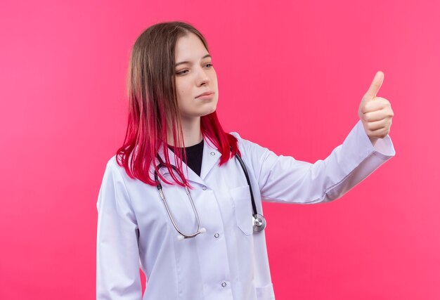 Auf der Seite junge Ärztin Mädchen tragen Stethoskop medizinische Kleid ihren Daumen oben auf rosa isolierten Hintergrund