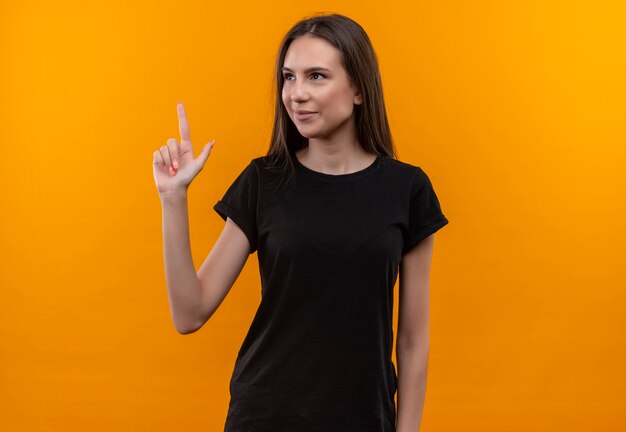 Auf der Seite lächelndes junges kaukasisches Mädchen, das schwarzes T-Shirt trägt, zeigt nach oben mit Finger auf isolierte orange Wand