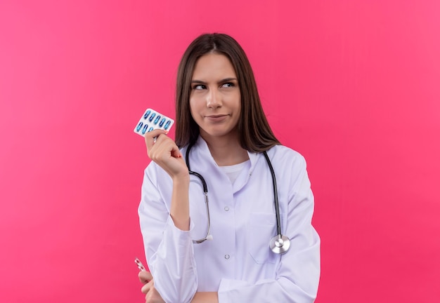 Auf der Seite verwirrte junge Ärztin, die das medizinische Kleid des Stethoskops hält, das Pillen auf isolierter rosa Wand hält