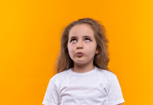 Auf der Suche nach einem kleinen Schulmädchen, das ein weißes T-Shirt trägt und eine Kussgeste auf einem isolierten orangefarbenen Hintergrund zeigt