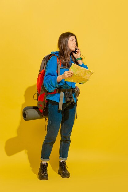 Auf der Suche nach Weg mit Karte. Porträt eines fröhlichen jungen kaukasischen Touristenmädchens mit Tasche und Fernglas lokalisiert auf gelbem Studiohintergrund. Vorbereitung auf Reisen. Resort, menschliche Gefühle, Urlaub.