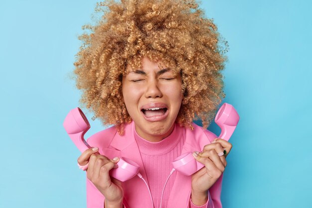 Aufgebrachte, weinende, lockige Frau verwendet ein Retro-Handy-Telefon, fühlt sich traurig, drückt negative Emotionen aus, trägt eine rosa Jacke, verwendet ein Retro-Objekt, das über blauem Hintergrund isoliert ist, kann nicht mit Verwandten sprechen