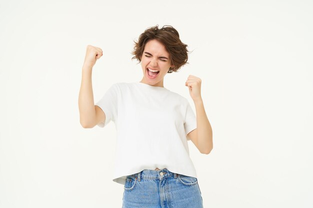 Aufgeregt brünette Studentin gewinnt den Preis und feiert den Sieg triumphierend im weißen T-Shirt stehend