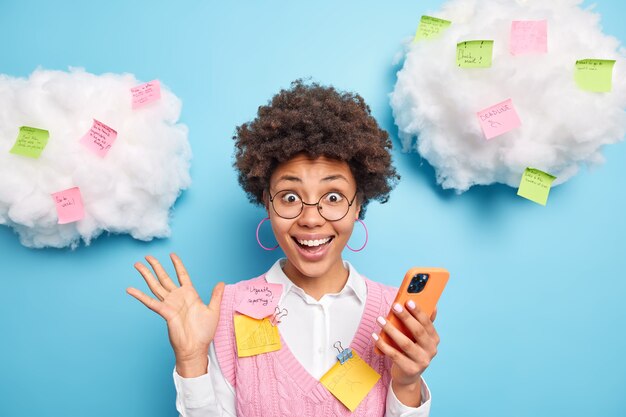 Aufgeregt fröhlich fleißig Student hält Smartphone findet heraus, hervorragende Ergebnisse der bestandenen Prüfung lächelt breit posiert gegen blaue Wand mit geklebten bunten Aufklebern auf Wolken