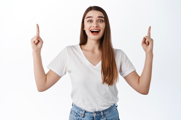 Aufgeregt lächelnde Studentin, die mit den Fingern nach oben zeigt, Promo-Deal zeigt, Werbebanner oben, gegen weiße Wand stehend