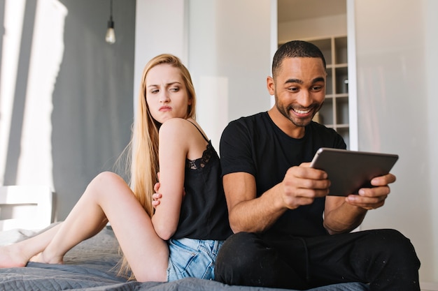 Aufgeregt lächelte gutaussehender Kerl, der im Internet-Tablet auf dem Bett in der Nähe von verärgerter wütender junger hübscher Frau mit langen blonden Haaren surft, die ihn ansieht. Spaß haben, Beziehung, zu Hause