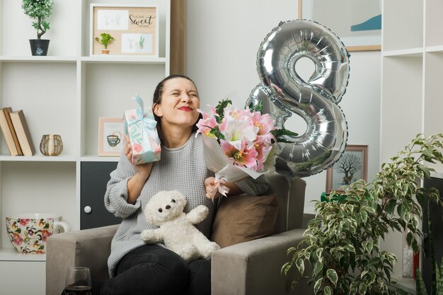 Aufgeregt mit geschlossenen Augen schönes Mädchen am glücklichen Frauentag, der Geschenk mit Blumenstrauß hält, der auf einem Sessel im Wohnzimmer sitzt