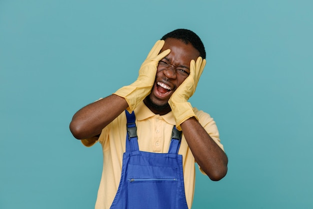 Aufgeregt packte Gesicht junge afroamerikanische Reinigungskraft in Uniform mit Handschuhen isoliert auf blauem Hintergrund