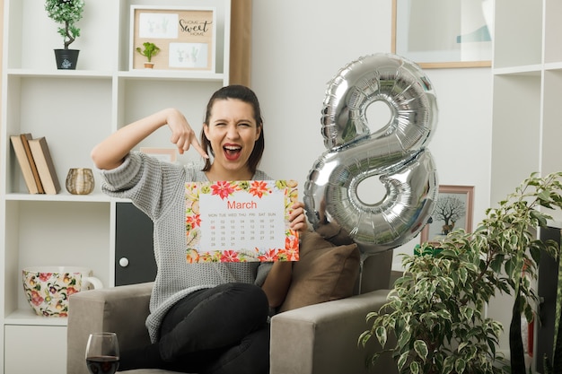 Aufgeregt schöne Frau am glücklichen Frauentag hält und zeigt auf den Kalender, der auf einem Sessel im Wohnzimmer sitzt