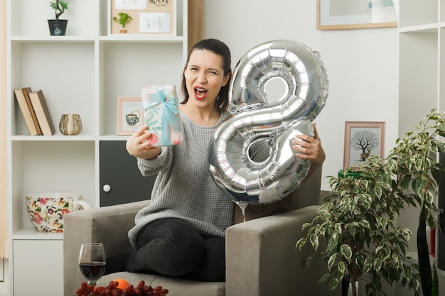 Aufgeregt schöne frau am glücklichen frauentag mit ballon nummer acht mit geschenk auf sessel im wohnzimmer sitzend Premium Fotos