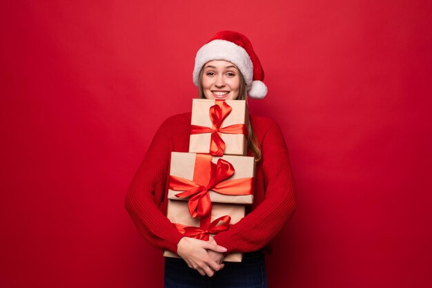 Aufgeregt überraschte Frau im roten Weihnachtsmann-Outfit, die Stapelgeschenke isoliert auf der roten Wand hält