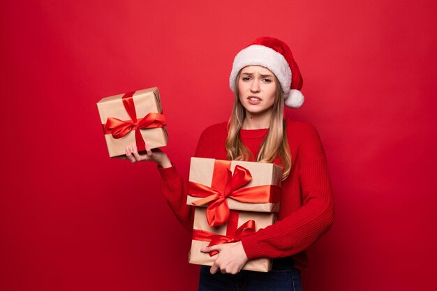 Aufgeregt überraschte Frau im roten Weihnachtsmann-Outfit, die Stapelgeschenke isoliert auf der roten Wand hält