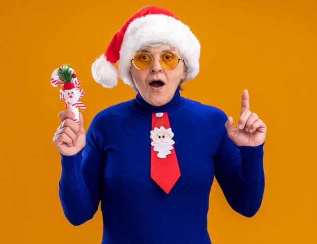 aufgeregte ältere Frau in Sonnenbrille mit Weihnachtsmütze und Weihnachtskrawatte hält Zuckerstange und zeigt lokalisiert auf orangefarbenem Hintergrund mit Kopienraum