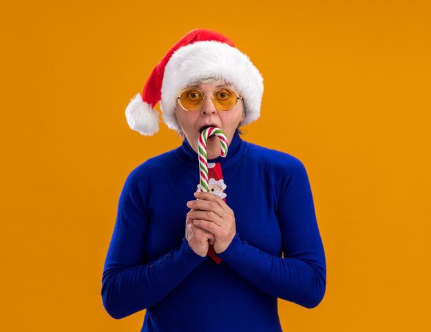 Aufgeregte ältere frau in sonnenbrille mit weihnachtsmütze und weihnachtsmann-krawatte, die zuckerstange einzeln auf orangefarbener wand mit kopienraum isst