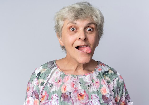 Aufgeregte ältere Frau streckt Zunge heraus und sieht isoliert auf weißer Wand aus
