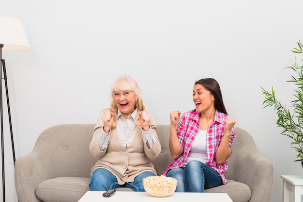Kostenloses Foto aufgeregte ältere mutter- und erwachsentochter, die auf dem sofa sitzt, das ihre faust beim fernsehen zusammenpreßt