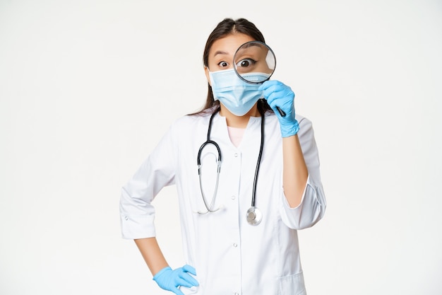 Aufgeregte asiatische medizinische arbeiterin schaut durch lupe überrascht beim tragen von...