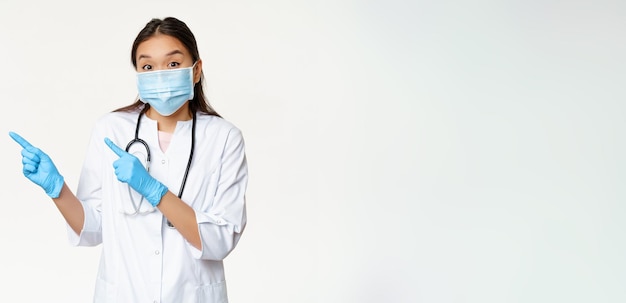 Aufgeregte asiatische Ärztin weist nach links und trägt eine medizinische Maske als präventive Maßnahmen