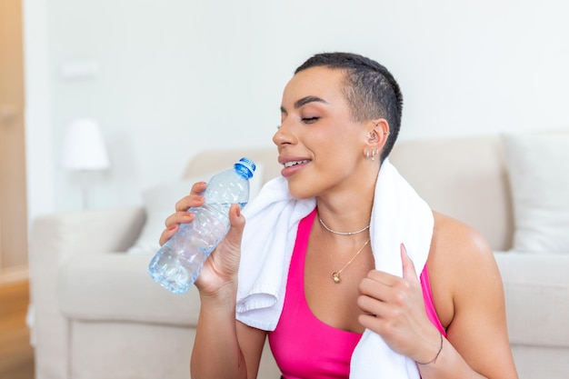 Aufgeregte athletische schwarze Frau in Sportkleidung, die lachend auf einer Yogamatte auf dem Boden sitzt und eine Flasche Wasser mit einem Handtuch auf ihren Schultern hält