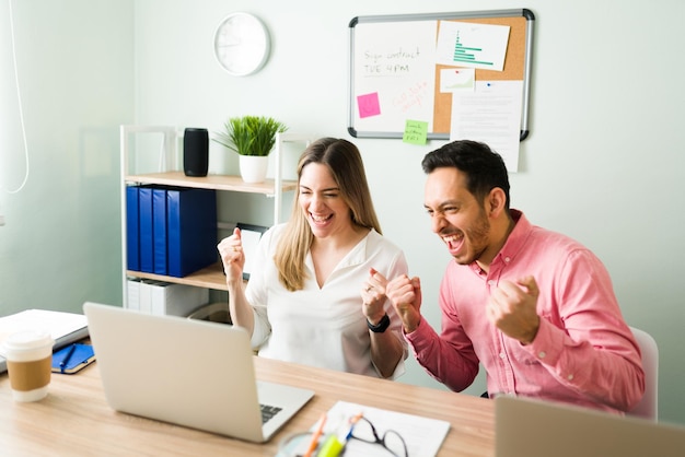 Aufgeregte Berufskollegen schreien und jubeln während eines Online-Meetings im Büro. Geschäftspartner feiern, nachdem sie einen Arbeitsvertrag bei einem Videoanruf auf ihrem Laptop abgeschlossen haben