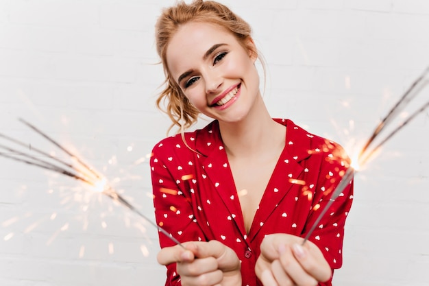 Aufgeregte Dame mit trendigem Make-up, das Wunderkerzen auf weißer Wand hält. hübsches glückliches Mädchen mit blonden Haaren, die mit bengalischen Lichtern aufwerfen.