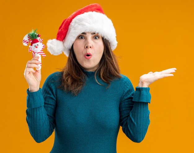 aufgeregte erwachsene kaukasische Frau mit Weihnachtsmütze hält Zuckerstange und hält Hand offen lokalisiert auf orange Hintergrund mit Kopienraum