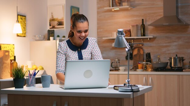 Aufgeregte Frau fühlt sich ekstatisch beim Lesen großartiger Online-Nachrichten auf dem Laptop, der von der heimischen Küche aus arbeitet. Glücklicher Mitarbeiter, der moderne drahtlose Technologie verwendet, Überstunden beim Schreiben und Suchen macht