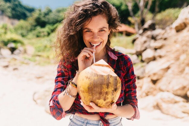 Aufgeregte glückliche Frau gekleidete Shorts und helles Hemd sitzen am Strand mit Kokosnusscocktail