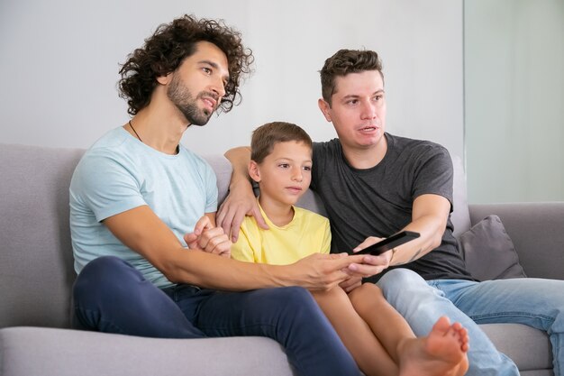 Aufgeregte homosexuelle Väter und Sohn sehen zu Hause Fernsehsendungen, sitzen auf der Couch im Wohnzimmer, umarmen sich, benutzen die Fernbedienung und schauen weg. Familien- und Home-Entertainment-Konzept