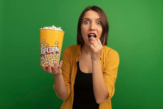 Aufgeregte hübsche kaukasische Frau isst und hält Eimer Popcorn auf Grün