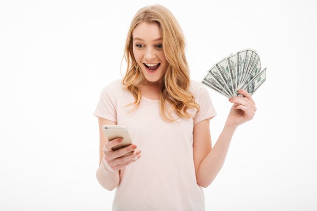 Aufgeregte junge Dame, die Geld unter Verwendung des Handys hält.